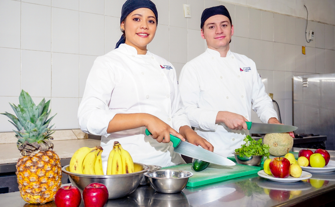 Dos estudiantes de gastronomía posan para una foto cortando fruta